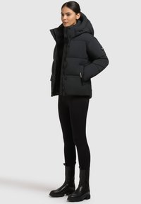 Čierna puffová bunda s kapucňou, prešívaný dizajn, predné gombíky a bočné vrecká. Nosená cez čierny outfit a skombinovaná s čiernymi členkovými čižmami.