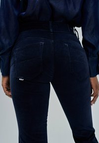 Persona vestida con jeans de tiro alto en azul oscuro y camisa a juego en azul oscuro, con las manos relajadas a los lados, mostrada desde la cintura hasta la mitad del muslo.