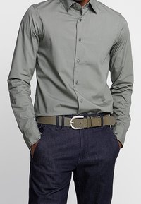 Jasnoszara koszula z długim rękawem, zestawiona z ciemnoniebieskimi dżinsami i plecionym oliwkowozielonym paskiem z srebrną klamrą.