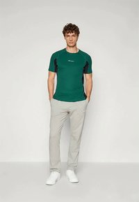 Tricou sportiv verde cu accente negre, mâneci scurte și logo; asociat cu pantaloni de trening gri deschis și adidași albi.