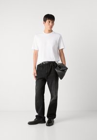 Weißes Baumwoll-T-Shirt mit eingeprägtem Logo, kombiniert mit schwarzen Jeans. Trägt eine schwarze Tasche und trägt schwarze Schuhe. Schlichtes Design.