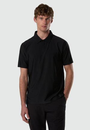 Giovane uomo con capelli ricci che indossa una polo nera a maniche corte e pantaloni neri, in piedi con una mano in tasca.