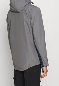Szara wodoodporna kurtka z kapturem, z kontrastowym granatowym bocznym panelem, gładką fakturą i elastycznymi mankietami. Zaprojektowana do użytku na zewnątrz.