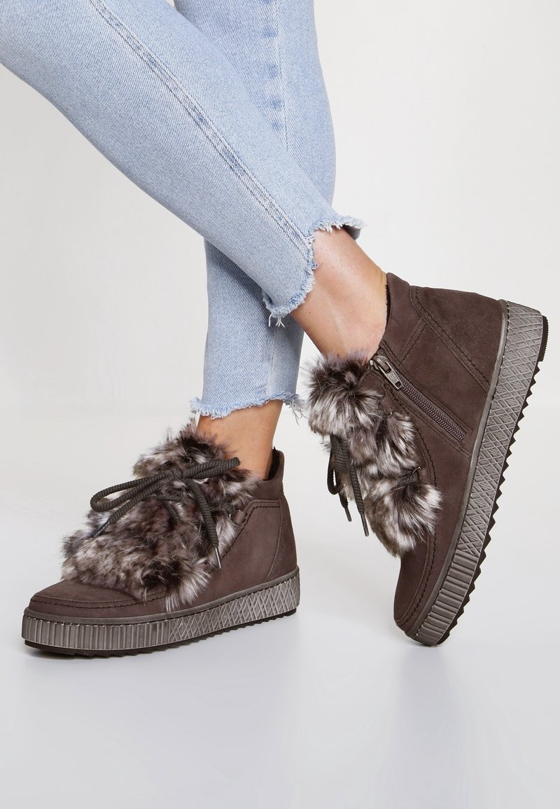 Zapatillas altas de ante marrón con acentos de pelaje sintético, suela de goma texturizada y diseño de cordones. Unos jeans de mezclilla deshilachados complementan el calzado.