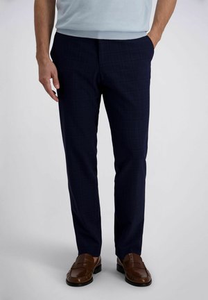 Homme portant un pantalon de costume à carreaux bleu marine, une chemise bleu clair rentrée, et des mocassins en cuir marron, debout avec une main dans la poche.