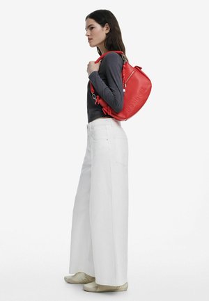 Sac à dos en cuir rouge de forme arrondie, avec une surface texturée et des détails de marque. Le modèle porte un pantalon large blanc et des chaussures claires.