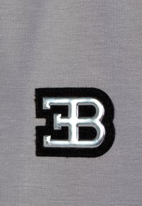 Grauener Stoff mit einem auffälligen schwarzen Filzpatch und einem silbernen Metall-"EB"-Logo, das eine glatte Textur und klare Buchstaben aufweist.