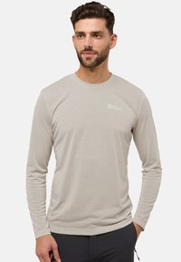 Mann mit kurzem dunklem Haar und Bart, der ein beiges langärmliges Jack Wolfskin-Shirt und eine dunkle Hose trägt und mit den Händen an den Seiten steht.