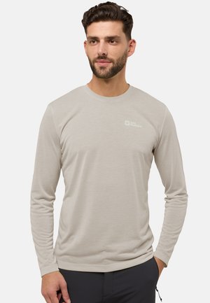 Man met kort donker haar en een baard, gekleed in een beige Jack Wolfskin overhemd met lange mouwen en donkere broek, staand met de handen langs zijn zij.