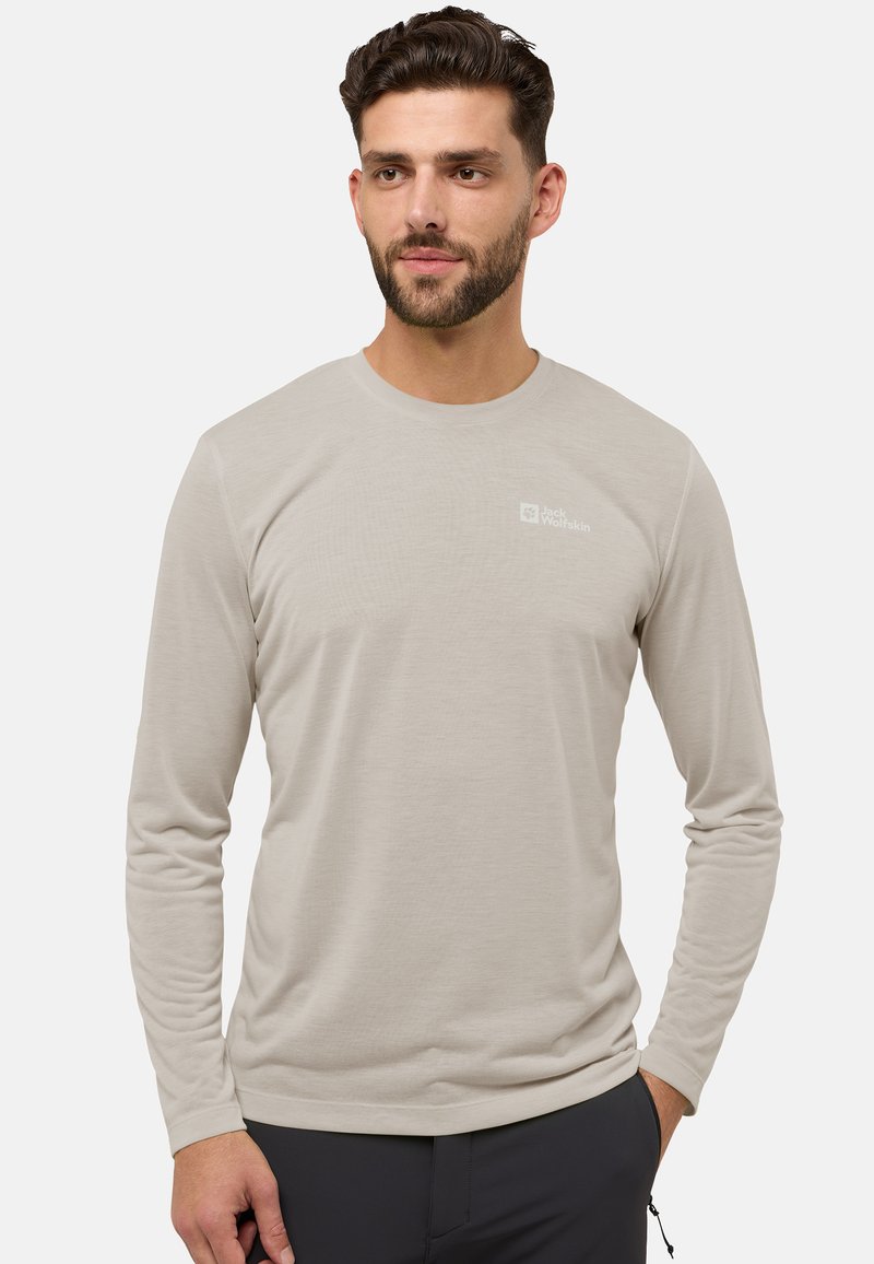 Mann mit kurzem dunklem Haar und Bart, der ein beiges langärmliges Jack Wolfskin-Shirt und eine dunkle Hose trägt und mit den Händen an den Seiten steht.