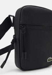 Black textured crossbody bag made from synthetic material. Features a zippered pocket, adjustable strap, and a small embroidered logo.