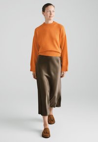 Pomarańczowy sweter na drutach, oliwkowa spódnica z satyny oraz brązowe zamszowe wsuwane buty z frędzlami. Prosty fason o luźnym kroju.