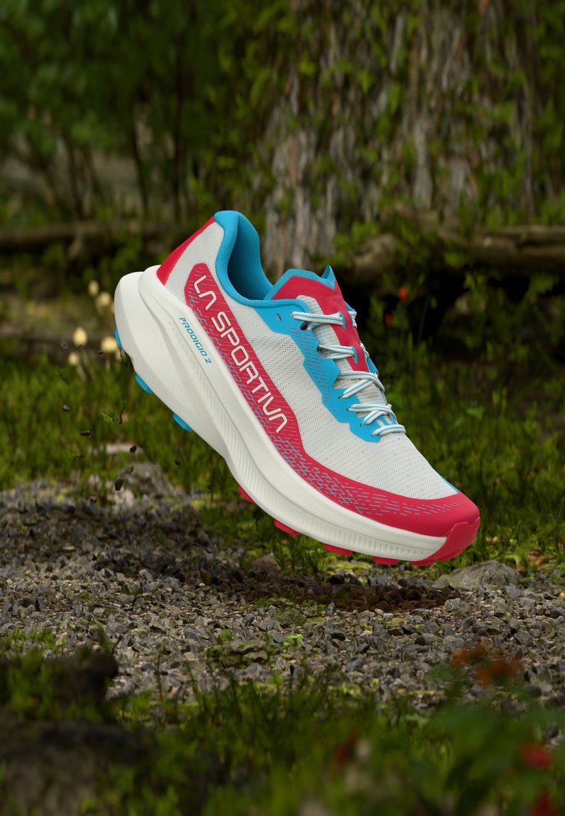 Pantof de alergare montană alb, roșu și albastru cu sigla La Sportiva, suspendat deasupra unui teren stâncos în aer liber, cu vegetație în fundal.