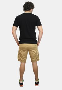 Maglietta nera a maniche corte, pantaloni cargo beige con tasche laterali e scarpe nere con accenti arancioni e verdi; vista da dietro.