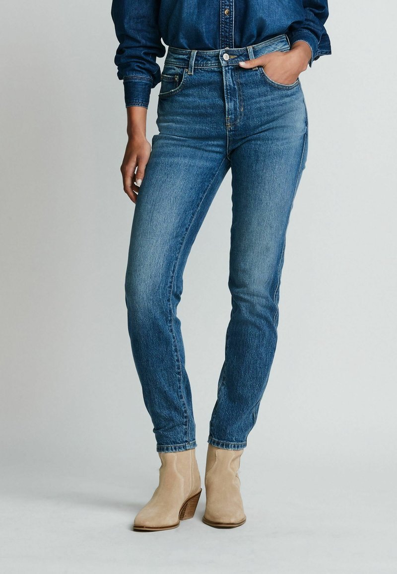 Personne portant un jean taille haute bleu, une chemise en denim bleu foncé et des bottines en daim beige sur un fond uni clair.