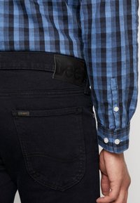 Jean noir à finition texturée, avec un patch logo à la taille et une poche arrière. Chemise à manches longues bleu à carreaux avec poignets boutonnés.