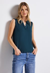 Turkusowa bluzka bez rękawów z dekoltem w kształcie litery V, wykonana z miękkiego materiału, o luźnym kroju, zestawiona z jasnoniebieskimi dżinsami. Minimalne szwy, gładka tekstura.