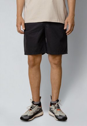 Mężczyzna w czarnych szortach i szarych butach sportowych z pomarańczowymi i czarnymi akcentami stoi na jednolitym tle.