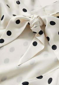Tissu blanc avec de grands pois noirs, présentant un détail de cravate. La texture est lisse et légère, avec une apparence légèrement transparente.
