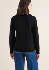 Mujer de espaldas vestida con una camiseta negra de manga larga y jeans azules, con cabello castaño hasta los hombros, frente a un fondo neutro.
