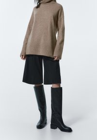 Osoba ubrana w luźny beżowy sweter z golfem, czarne szerokie szorty do kolan oraz czarne skórzane kozaki do kolan na białym tle.