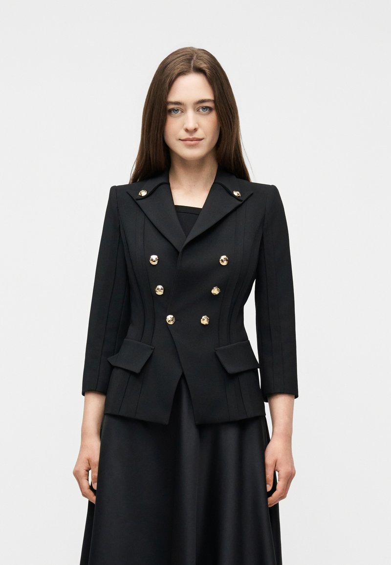 Γυναίκα με μακριά καστανά μαλλιά, φοράει εφαρμοστή μαύρη σακάκι με χρυσά κουμπιά και 3/4 μανίκια, στέκεται μπροστά σε ένα απλό ανοιχτό φόντο.