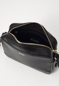 Borsa a tracolla nera in pelle con chiusura zip, hardware dorato e interno foderato, dotato di tasca laterale e dettaglio del logo.