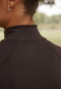 Top sport negru cu guler înalt, având un logo „ACTIVE” în relief pe spate. Textură netedă cu detalii vizibile de cusătură.