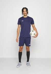 Tamsiai mėlynas sportinis marškinėlis ir šortai su balta logotipu, derinami su juodomis kojinėmis ir pilkais sportbačiais. Laiko daugiakampį futbolo kamuolį.