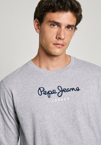 Szürke, hosszú ujjú pamut póló, amelyen sötétkék "Pepe Jeans London" felirat található a mellkason. Kerek nyakkivágás, laza fazon.