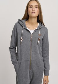 Tuta grigia con zip e cappuccio, dotata di zip frontale, cordino decorativo e tasche laterali; realizzata in un morbido tessuto testurizzato.