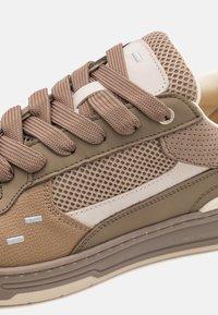 Gros plan sur une basket beige et marron avec un mesh texturé, des empiècements en daim et cuir, des lacets épais couleur fauve et des détails perforés près du talon.