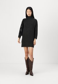 Robe en tricot noir avec un col haut et des manches longues, accompagnée de bottes de cow-boy marron avec des détails texturés. Porte un sac à main noir.