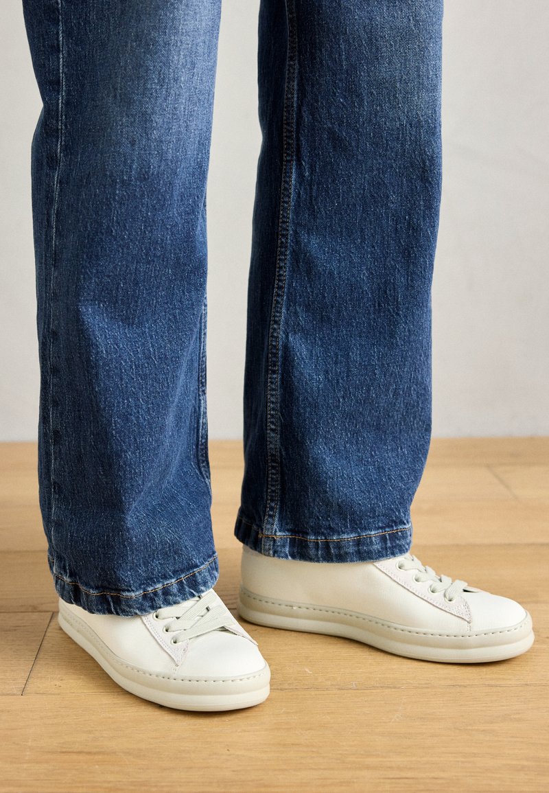 Weiße Lederturnschuhe mit runden Zehen und flachen Schnürsenkeln, kombiniert mit lockeren blauen Jeans, die auf einem Holzboden ruhen.
