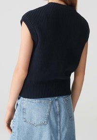 Kvinna i en ärmlös svart stickad tröja med ribbad nederkant, tillsammans med ljusblå högmidjade jeans, sett bakifrån.