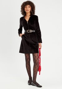 Robe en velours côtelé noire avec une fermeture éclair, taille cintrée par une large ceinture noire avec une boucle en or, associée à des collants à pois et des chaussures noires.