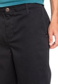 Černé kalhoty z pevné látky s hladkým povrchem, vybavené zapínáním na knofl íky a bočními kapsami, s malým logem jako doplňkem.