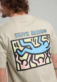 Camiseta de algodón beige con un gráfico colorido de Keith Haring en la parte posterior, con líneas y formas llamativas en azul, amarillo y rosa.