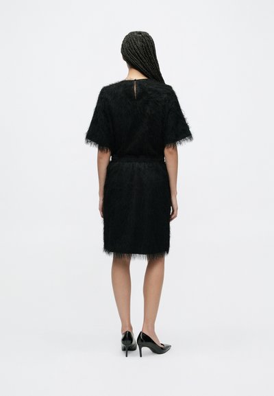 Robe noire à texture duveteuse, manches courtes, taille ajustée et ourlet jusqu'aux genoux, associée à des escarpins noirs.