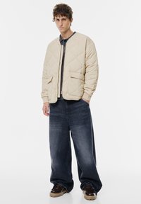 Beige quiltet bomberjakke med ribbede manchetter, parret med mørke, posede denimjeans og brune snørebåndssko. Enkel, funktionel design.