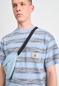 Lys blåstripet skjorte med korte ermer og brystlomme, samt en fremtredende Carhartt-logo; kombinert med en lys blå crossover-veske.