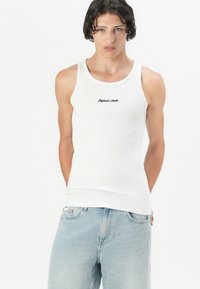 Hvit ribbestrikket tank topp med rund hals, med teksten "Originals Studio" i svart kursiv på fronten, kombinert med lyseblå denimjeans.