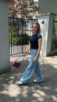 Rød skuldertaske med et struktureret design, båret med en tætsiddende marineblå bluse og løse, lyseblå denimjeans. Modellen har hvide sneakers med sorte striber på.