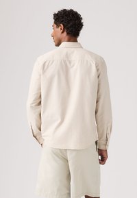 Homme aux cheveux bouclés portant une chemise à manches longues beige clair et un short assorti, debout de dos sur un fond uni.