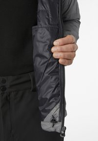 Donkergrijze geïsoleerde jas met een ritssluiting en een binnenzak. Hand die de rits vasthoudt, wat een gladde, glanzende textuur toont.
