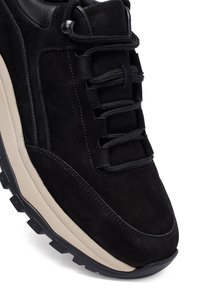 Sneaker nera in suede con tomaia strutturata, lacci piatti neri e suola intermedia beige. Presenta un robusto battistrada in gomma con un motivo di trazione.