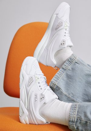 Las zapatillas Fila blancas presentan un diseño elegante con cuero texturizado, perforaciones, acentos verdes y una suela acolchada. Se llevan con jeans azules y calcetines blancos.
