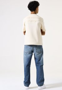 Korte mouwen crèmekleurig sweatshirt met reliëftekst "THE FUTURE EDITION" op de rug, gecombineerd met lichtblauwe denim jeans en witte sneakers.
