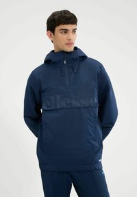 Marinefarbener Windbreaker mit Kapuze, Kordelzug und einer Fronttasche. Hergestellt aus einem glatten, leichten Material und mit einem dezenten Logo auf der Brust.