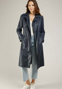 Manteau trench en cuir bleu marine avec un design croisé, de larges revers et des poches latérales ; associé à un jean clair et des baskets blanches.